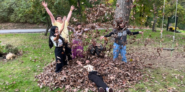 A diverse group of Rock Point School students play in a pile of leaves on campus in Burlington, Vermont. Students are rolling in the leaves, throwing leaves in the air, and having fun together.